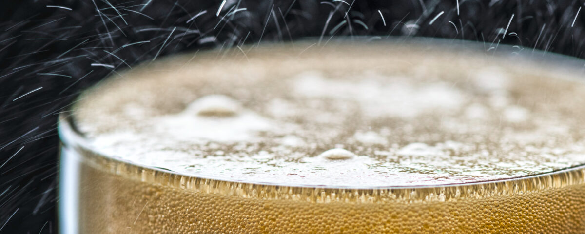 Imagem de um copo com líquido escuro e espuma na superfície, indicando um refrigerante ou bebida carbonatada. Pequenas bolhas são visíveis na espuma, enquanto gotículas e partículas de gás sobem do líquido, ilustrando o processo de carbonatação. A cena tem um fundo escuro que destaca o brilho das bolhas e a efervescência característica da carbonatação.