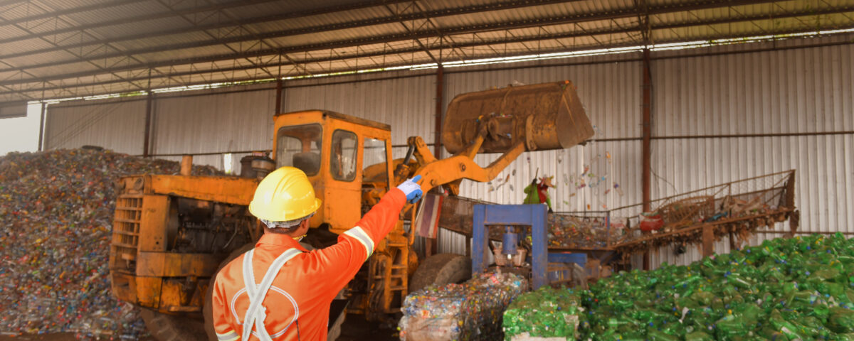 Um trabalhador, equipado com capacete amarelo e uniforme laranja de segurança, aponta para uma máquina escavadeira dentro de um galpão de triagem de resíduos. A máquina movimenta materiais recicláveis, principalmente garrafas plásticas, que estão acumuladas em grandes pilhas ao fundo e em fardos compactados à frente. O cenário representa a gestão de resíduos, destacando a importância da separação, processamento e reaproveitamento de materiais em uma operação industrial focada em sustentabilidade e economia circular