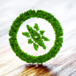 Um símbolo verde, feito com folhas e vegetação, representa um círculo com uma árvore estilizada no centro. Esse elemento visual remete à neutralidade de carbono, destacando práticas sustentáveis e o compromisso com a compensação de emissões de CO₂. A imagem sugere harmonia entre progresso e meio ambiente, simbolizando ações como reflorestamento, uso de fontes renováveis e a busca por um equilíbrio ecológico em prol da preservação do planeta.