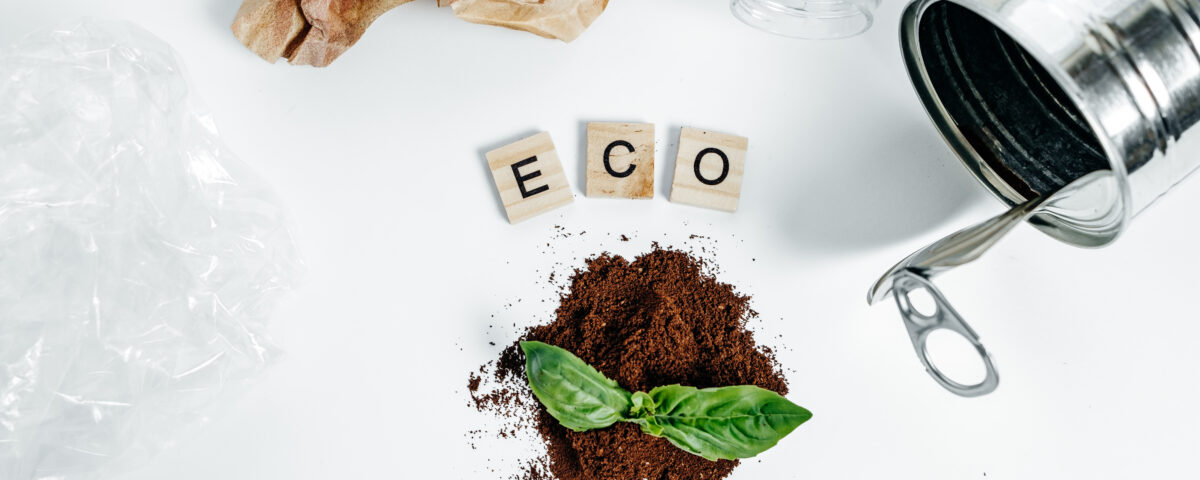 Imagem conceitual sobre biossoluções e sustentabilidade. No centro, a palavra 'ECO' é formada por três blocos de madeira, rodeada por elementos que remetem ao reaproveitamento e reciclagem: uma lata metálica aberta, um recipiente de plástico transparente, papel amassado e um pedaço de plástico. Em primeiro plano, uma pequena pilha de borra de café usada serve de substrato para uma folha verde, simbolizando o reaproveitamento de resíduos orgânicos como solução ecológica.