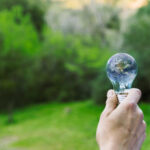 Mão segurando uma lâmpada transparente, na qual a imagem do planeta Terra está refletida, em um cenário natural com árvores e vegetação verde ao fundo. A composição simboliza a conexão entre sustentabilidade e energia verde, destacando a importância de soluções energéticas ecológicas para o futuro do planeta.