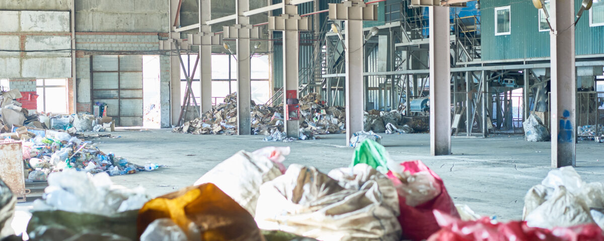Imagem de um galpão industrial espaçoso, com estrutura metálica visível e luz natural entrando pelas janelas ao fundo. O local contém pilhas de resíduos industriais espalhados, incluindo sacos plásticos coloridos e materiais descartados, como papelão e plástico. Ao fundo, há equipamentos metálicos e estruturas indicando um possível sistema de triagem ou processamento de resíduos. A cena reflete um ambiente relacionado à gestão de resíduos industriais, evidenciando a necessidade de organização e processos adequados para lidar com o descarte e reciclagem desses materiais.