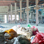 Imagem de um galpão industrial espaçoso, com estrutura metálica visível e luz natural entrando pelas janelas ao fundo. O local contém pilhas de resíduos industriais espalhados, incluindo sacos plásticos coloridos e materiais descartados, como papelão e plástico. Ao fundo, há equipamentos metálicos e estruturas indicando um possível sistema de triagem ou processamento de resíduos. A cena reflete um ambiente relacionado à gestão de resíduos industriais, evidenciando a necessidade de organização e processos adequados para lidar com o descarte e reciclagem desses materiais.