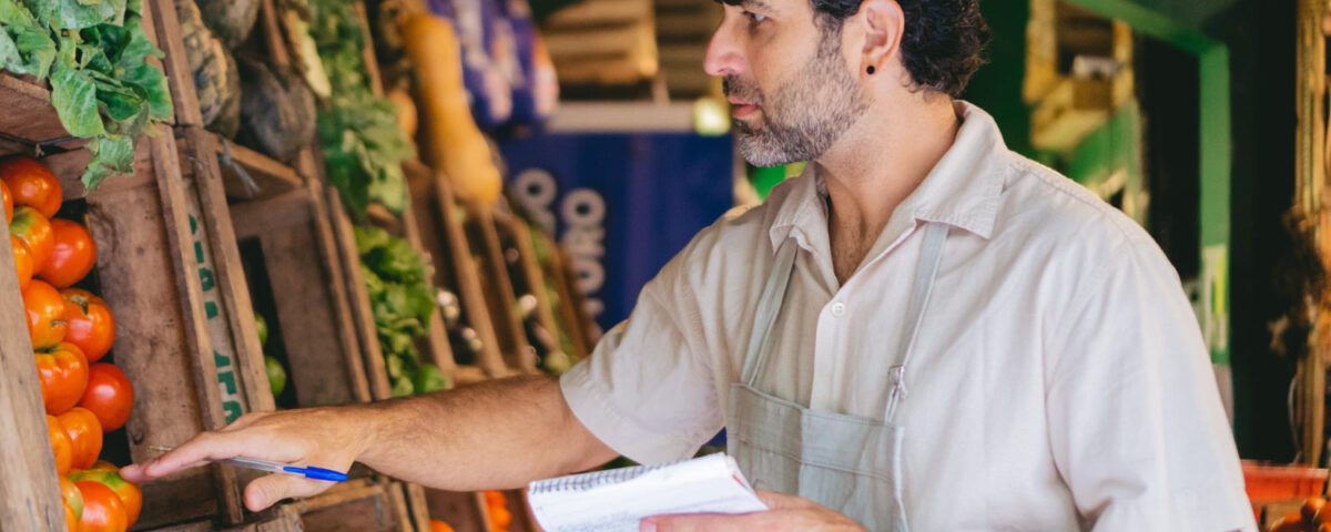 A imagem mostra um homem de cabelos cacheados e barba analisando cuidadosamente tomates frescos em uma feira ou mercado. Ele veste um avental claro e segura um caderno e uma caneta, sugerindo que está realizando um controle de estoque ou avaliação da qualidade dos produtos. Ao redor, prateleiras de madeira repletas de hortaliças e legumes indicam a variedade de alimentos disponíveis. O cenário remete à crescente alta demanda por alimentos, exigindo um acompanhamento rigoroso para garantir abastecimento e qualidade.