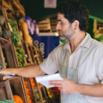 A imagem mostra um homem de cabelos cacheados e barba analisando cuidadosamente tomates frescos em uma feira ou mercado. Ele veste um avental claro e segura um caderno e uma caneta, sugerindo que está realizando um controle de estoque ou avaliação da qualidade dos produtos. Ao redor, prateleiras de madeira repletas de hortaliças e legumes indicam a variedade de alimentos disponíveis. O cenário remete à crescente alta demanda por alimentos, exigindo um acompanhamento rigoroso para garantir abastecimento e qualidade.