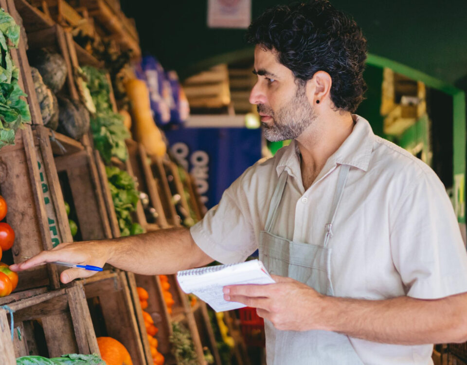 A imagem mostra um homem de cabelos cacheados e barba analisando cuidadosamente tomates frescos em uma feira ou mercado. Ele veste um avental claro e segura um caderno e uma caneta, sugerindo que está realizando um controle de estoque ou avaliação da qualidade dos produtos. Ao redor, prateleiras de madeira repletas de hortaliças e legumes indicam a variedade de alimentos disponíveis. O cenário remete à crescente alta demanda por alimentos, exigindo um acompanhamento rigoroso para garantir abastecimento e qualidade.